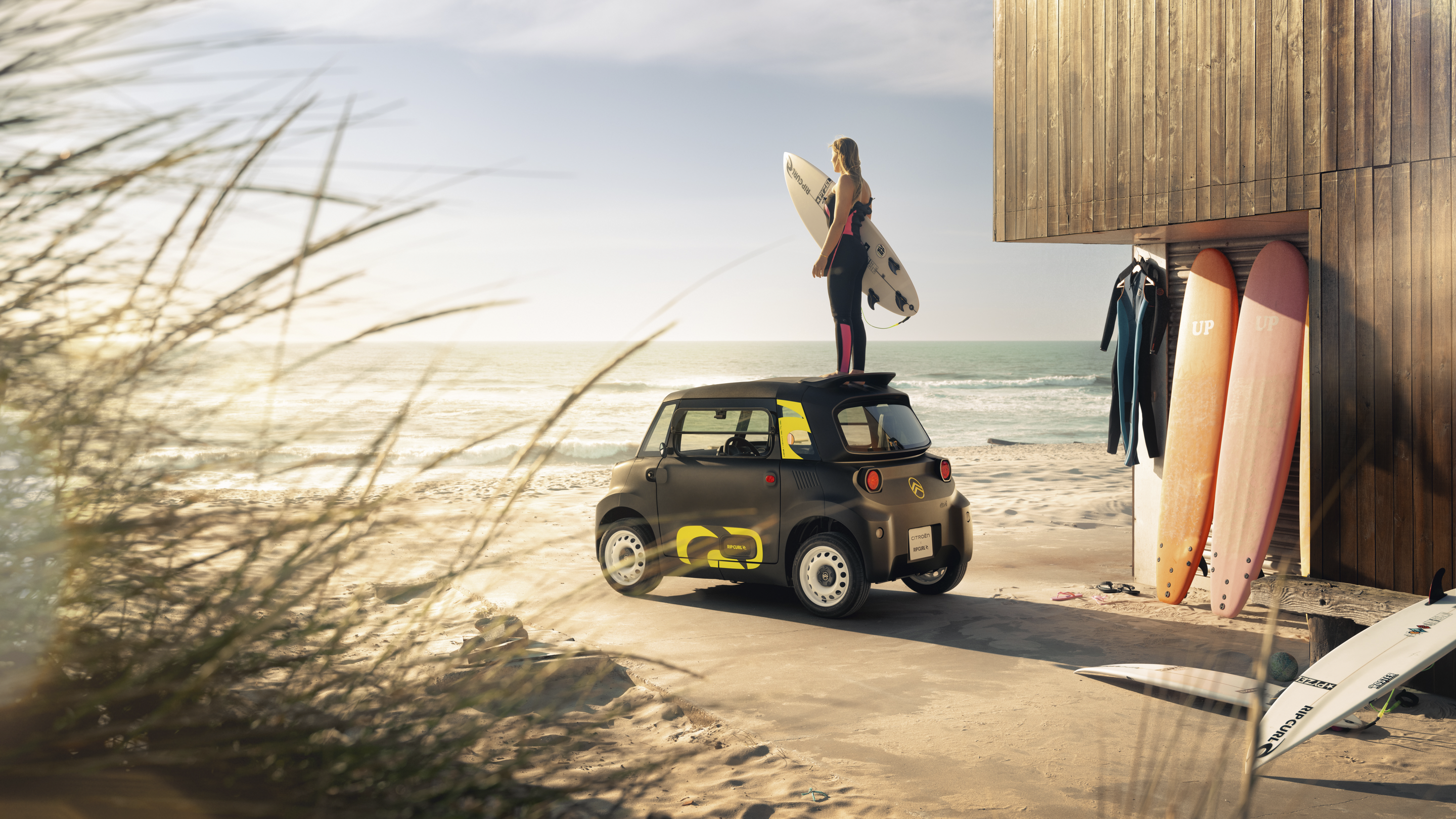 Achteraanzicht in lifestyle‑setting aan het strand van de Citroën Ami x Rip Curl, met surf‑ en kustgevoel voor stedelijke mobiliteit.